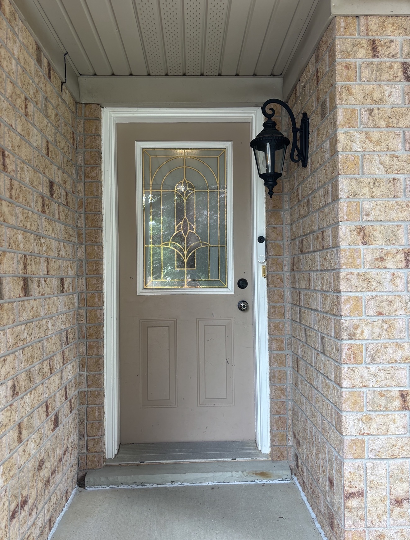 Before — beige steel entry door with ornate leaded-glass insert, two lower recessed panels, original white trim and hardware, set into a light brick alcove