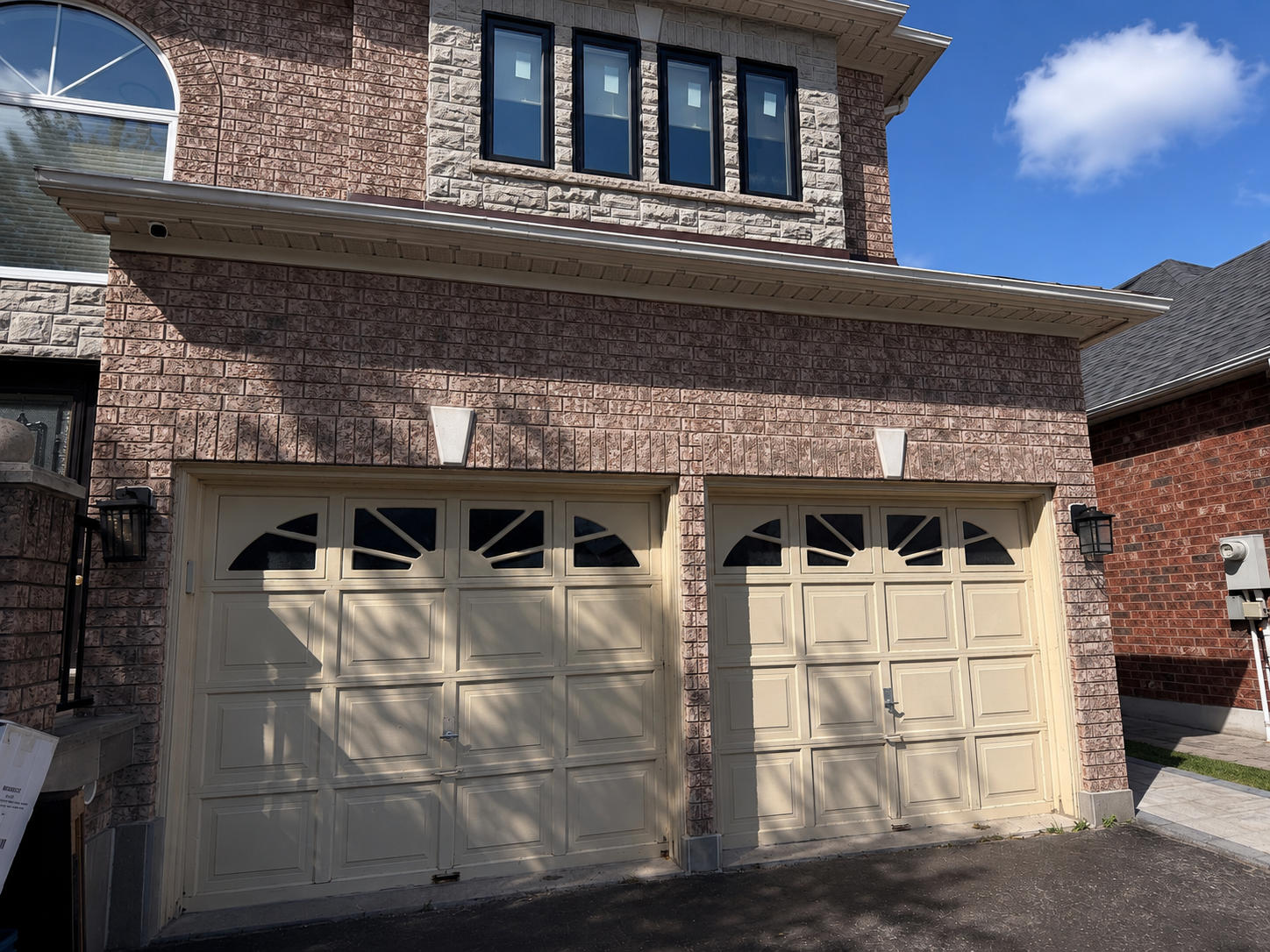 Before — dated cream raised-panel garage doors with arched sunburst window pattern, worn finish, set into a stone and brick two-storey facade
